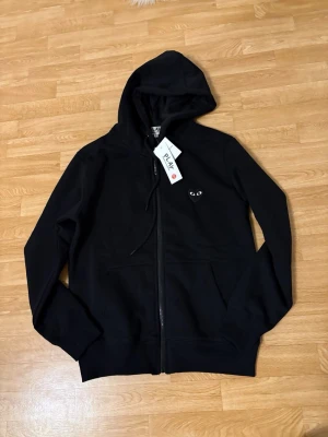 Svart hoodie från Comme des Garçons Play - Svart hoodie med dragkedja från Comme des Garçons Play. Framtill finns det klassiska hjärtloggan med ögon och på ryggen ett stort svart hjärta med gula ögon. Mjuk bomullskänsla, huva med dragsko och fickor framtill. Perfekt för en stilren streetlook.