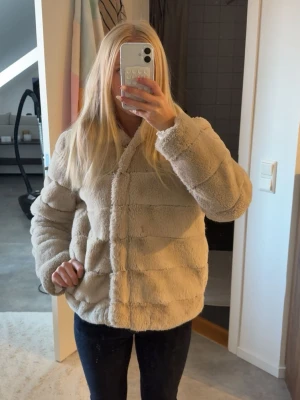 Beige teddyjacka med huva - Supermjuk och fluffig teddyjacka i beige med stora horisontella sömmar och generös huva. Jackan har en avslappnad passform och stängs framtill. Perfekt för dig som vill ha en cozy vibe och hålla dig varm under kyliga dagar.