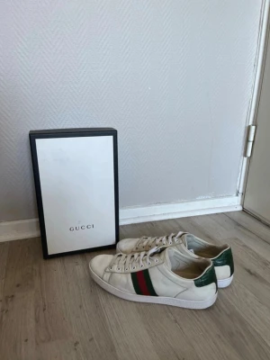 Gucci Ace skor - Gucci Ace sneakers i vitt läder med klassiska gröna och röda ränder på sidorna samt grön häl med metallic-finish. Låga sneakers med vit gummisula och snörning framtill. En ikonisk modell som ger en lyxig touch till din outfit.