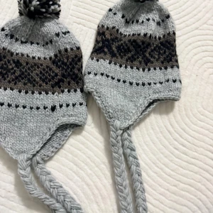Stickade öronlapparmössor med tofs - En för 50 Två stickade mössor i ljusgrått med brunt och svart mönster, öronlappar och flätade snören. Toppad med en fluffig tofs. Perfekt för kalla dagar och vinteraktiviteter. Jag har tre av de