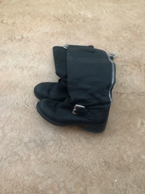 Svarta boots med dragkedja och spänne - Säljer ett par svarta boots i skinn med grov dragkedja längs skaftet och ett coolt spänne vid ankeln. Bootsens design är lite slouchy och de har en robust sula. Perfekta för en edgy streetstil. 