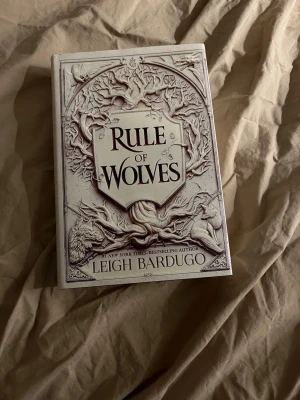 Rule of Wolves av Leigh Bardugo - Fantastisk roman på engelska, 'Rule of Wolves' av Leigh Bardugo. Omslaget är detaljerat med grenar och vargar i relief, i grå och vita toner. Perfekt för dig som gillar fantasy och Grishaverse-världen. Hardback cover. 