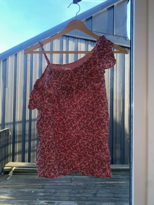 Rosa blommig offshoulder topp - Söt rosa offshoulder topp med blommigt mönster och volangdetalj längs halsringningen. Toppen har en axelbandslös sida och ett smalt axelband på andra sidan. Perfekt för dig som gillar en romantisk och trendig stil.