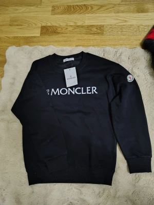 Svart Moncler sweatshirt - Svart sweatshirt från Moncler med vit logga tryckt över bröstet och klassisk Moncler-patch på ärmen. Tröjan har rund halsringning, ribbade muddar och är tillverkad i mjukt bomullsmaterial. Perfekt för en clean och stilren look.