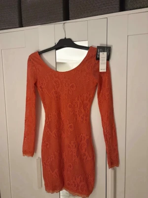 Korallfärgad spetsklänning från Nelly - Snygg kort spetsklänning i korall från Nelly med lång ärm och djup snörad rygg. Klänningen har figurnära passform och vackert blommönster i spets. Perfekt för dig som vill sticka ut med en unik detalj på ryggen.