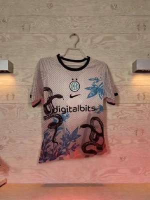 Inter Milan bortatröja Thuram #9 - Inter Milan bortatröja från Nike med Thuram och nummer 9 på ryggen. Tröjan är vit med svarta och blå detaljer, ormmönster och bladmönster framtill. Svart krage och ärmslut. Digitalbits sponsorlogga på bröstet. Perfekt för fotbollsfans!