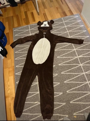 Brun björndräkt med huva - Mysig brun björndräkt i mjuk fleece med dragkedja framtill. Dräkten har en vit mage och huva med björnansikte och öron. Perfekt för maskerad eller cosplay när du vill vara extra gullig.