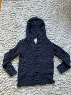 Mörkblå stickad cardigan med luva  - Mysig mörkblå stickad cardigan med grovt kabelstickat mönster och knäppning framtill. Storlek S men passar även XS.                                     