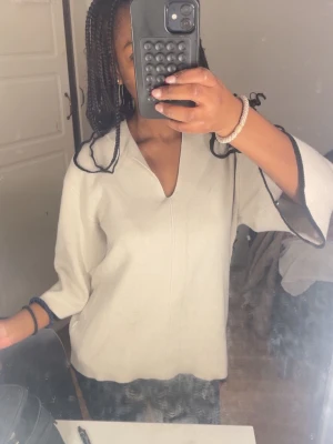 Vit oversized tröja med svarta detaljer - En vit oversized tröja med svarta kanter på ärmarna. Tröjan har en djup v-ringning och vida ärmar, vilket ger en avslappnad, elegant och trendig look.