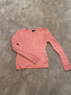 Rosa kabelstickad tröja Polo Ralph Lauren - Säljer en korallrosa kabelstickad tröja från Polo Ralph Lauren i mjuk pima cotton. Tröjan har rund halsringning, långa ärmar och den klassiska vita logon broderad på bröstet. Perfekt för dig som gillar en tidlös och fräsch stil. Skriv om ni vill ha mer bilder i bättre ljus så fixas det! (Pris går att diskuteras vid snabb affär). på etiketten står det large men den passar på S 