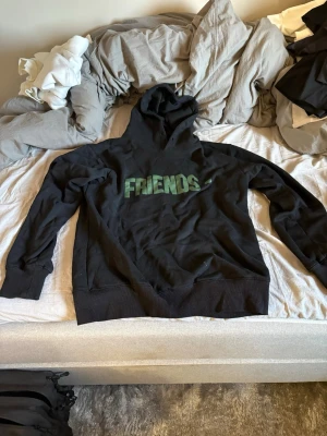 Svart Friends hoodie med tryck - Svart hoodie från VLONE med grön 'FRIENDS-' text på framsidan och stor grön V-logga med rödvit orm på ryggen. Klassisk passform med huva och muddar vid ärmslut och midja. Perfekt för dig som gillar streetwear och vill sticka ut.