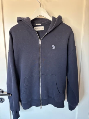 Mörkblå hoodie från Abercrombie & Fitch - Oversized mörkblå hoodie med dragkedja från Abercrombie & Fitch. Tillverkad i mjuk fleece, har huva, två stora fickor framtill och en liten broderad logga på bröstet. Perfekt för en chill och avslappnad stil. Förmodligen den skönaste tröjan jag haft men tyvärr för liten och orkade inte skicka tillbaka den så inte kommit till användning. 