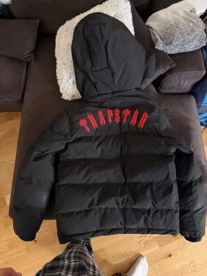 Trapstar svart pufferjacka med huva - Säljer en svart pufferjacka från Trapstar med stor huva och feta röda bokstäver på ryggen för ett billigt pris då jag inte använder den längre! Jackan har dragkedja framtill, två fickor och en rund Trapstar-logga på bröstet. Perfekt för kalla dagar när du vill se street ut.