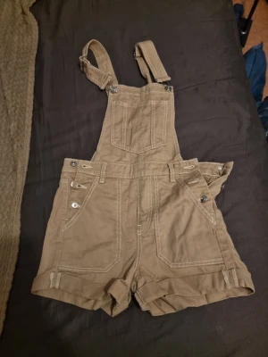 Beige hängselshorts från Divided - Säljer ett par snygga beige hängselshorts från Divided. Shortsen har justerbara axelband, flera fickor och coola knappdetaljer i silver. Tillverkade i ett slitstarkt bomullstyg med vita kontrastsömmar. Perfekta för en avslappnad och trendig look.