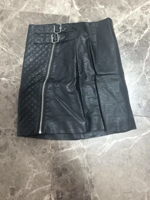 Svart skinnkjol med dragkedja och spännen - Cool svart kortkjol i skinnimitation med quiltad detalj på ena sidan, sned dragkedja framtill och två dekorativa spännen i midjan. Kjolen har en edgy vibe och passar perfekt till en trendig streetstyle-look.  Osäker på storleken men passar mig som är. Xs 