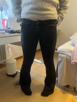 Svarta bootcut jeans, Joey - Säljer ett par svarta bootcut jeans från True Religion med låg midja och snygga detaljer på fickorna. Jeansen har knappar och dragkedja framtill samt dekorativa bakfickor med lock och knapp. Perfekta för dig som gillar en klassisk men trendig look. Midjemått: 76cm.      Innerbenslängd:79/80cm