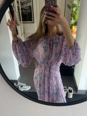 Blommig långklänning med volangärm - Säljer en romantisk långklänning i ljusa toner med blommigt mönster i rosa, blått och vitt. Klänningen har voluminösa långärmar, knyt i midjan och vid halsen samt volangdetaljer. Perfekt för sommarens alla tillfällen!