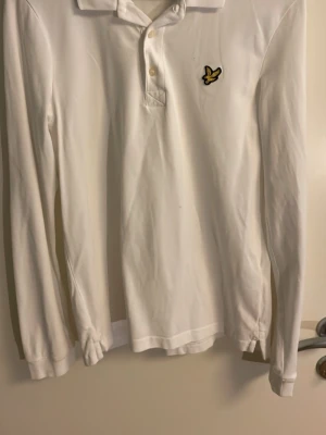 Vit långärmad skjorta från Lyle & Scott - Klassisk vit långärmad skjorta från Lyle & Scott med gul logga på bröstet. Skjortan har krage, knappar framtill och är tillverkad i mjuk bomull. Perfekt för en clean och stilren look. Perfekt vid golf med grabbarna nu till sommar