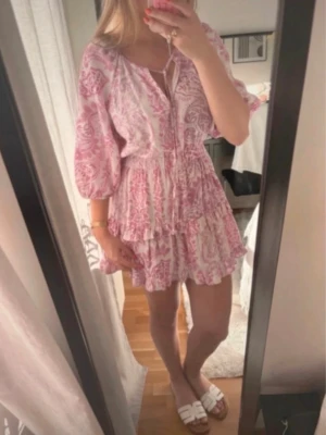  Trendig Rosa och vit kortklänning med volang - Söt kortklänning i rosa och vitt med blommigt mönster och volangdetaljer. Klänningen har trekvartsärmar, v-ringning med knyt och en luftig passform. Perfekt för varma dagar och somriga tillfällen.