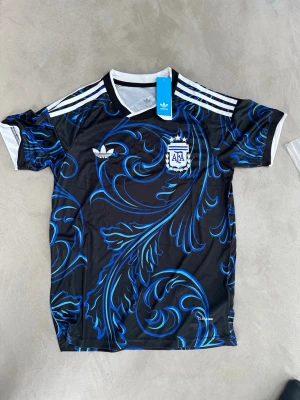 Argentina Adidas svart fotbollströja - Snygg svart fotbollströja från Adidas med blått och turkost mönster samt vita detaljer. Tröjan har AFA-logga på bröstet och klassiska Adidas-ränder på axlarna. Tillverkad i lätt och ventilerande material, perfekt för träning eller match.