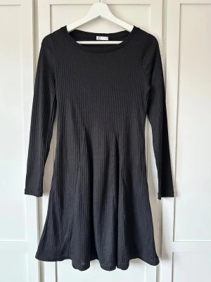 Svart ribbad klänning med lång ärm storlek S - En svart, ribbad klänning med lång ärm och rund halsringning. Modellen är kort och har en lätt utställd kjol som ger en snygg siluett. Perfekt för dig som gillar enkel och stilren design. Använd max 1-2 ggr.