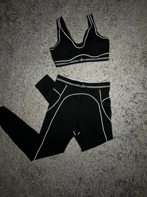 Svart set från Alo med vita detaljer - Snyggt svart set från Alo med sport-bh och tights. Setet har vita kontrastsömmar och logga framtill på både överdel och underdel. Perfekt för dig som gillar en sportig och trendig look. Materialet är stretchigt och följsamt.