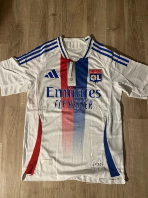 OL Lyon matchtröja Aouar - Snygg vit matchtröja från Olympique Lyonnais med Aouar och nummer 8 på ryggen. Tröjan har blå och röda detaljer, klubbmärke på bröstet och Adidas-logga. Tillverkad i lätt och ventilerande material med HEAT.RDY-teknologi. Perfekt för fotbollsträning eller support. HELT NY