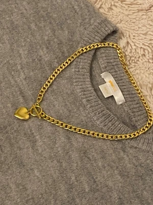 Guldfärgat halsband med hjärthänge - Stilrent halsband i guldfärgad rostfritt med en chunky kedja och ett hjärtformat hänge. Halsbandet har ett snyggt T-lås som detalj. Perfekt accessoar för att lyfta din outfit med en trendig touch.