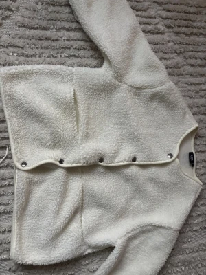 Vit teddykofta från 157 - Mysig vit cardiganjacka i teddy-material från 157. Jackan har rund halsringning, tryckknappar framtill och två stora fickor. Perfekt för lager på lager och ger en chill vibe till din outfit.