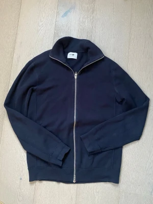Svart ziptröja från NN07 - Svart långärmad ziptröja från NN07 med hög krage och hel dragkedja framtill. Tröjan är i mjukt material och har ribbade muddar vid ärmslut och nederkant. Perfekt för en avslappnad och stilren look.