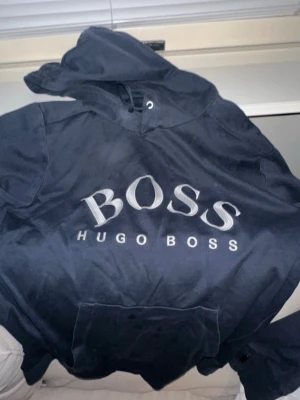 Svart hoodie från Hugo Boss - Snygg svart hoodie från Hugo Boss med stor logga broderad på bröstet. Klassisk modell med huva och känguruficka framtill. Perfekt för en chill och stilren look. Tjockt och mjukt material som känns skönt mot huden.