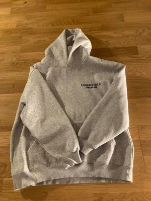 Grå Essentials Fear of God hoodie - Snygg grå hoodie från Essentials Fear of God med svart logga på bröstet. Klassisk passform med huva och känguruficka framtill. Tillverkad i mjukt material som är perfekt för en avslappnad streetwear-look.
