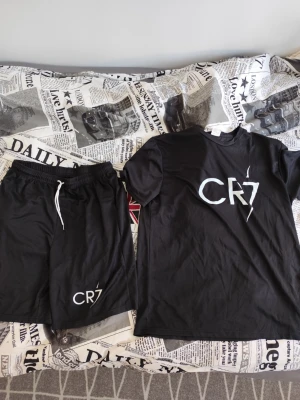 Svart CR7 set med t-shirt och shorts - Snyggt svart set från CR7 med matchande t-shirt och shorts. T-shirten har rund hals och tryckt CR7-logga framtill, shortsen har resår i midjan med vit dragsko och CR7-tryck på benet. Perfekt för en sportig och avslappnad look. Stor i storleken 