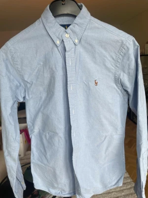 Ljusblå skjorta från Ralph Lauren - Klassisk somrig ljusblå skjorta från Ralph Lauren med button-down krage och broderad logga på bröstet. Skjortan har lång ärm och knäppning framtill. Perfekt för en clean och stilren look.