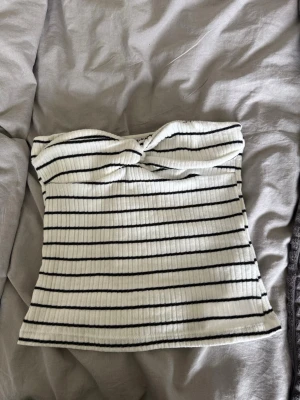 Randig ribbad tubtopp i vitt och svart - Snygg ribbad tubtopp i vitt med svarta horisontella ränder och twistad detalj framtill. Toppen är axelbandslös och har en tight passform som sitter snyggt på kroppen. Perfekt för en trendig och avslappnad look. NA-KD
