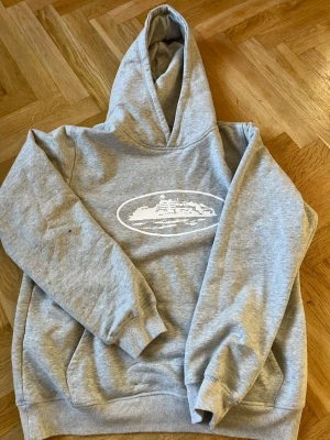 Grå hoodie med skepp-tryck - Grå hoodie med vit illustration av ett skepp på bröstet. Klassisk modell med huva och känguruficka framtill. Mjuk och skön bomullsblandning, perfekt för en avslappnad stil. Ribbade muddar vid ärmslut och nederkant.