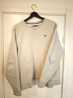 Grå sweatshirt från Lacoste Sport - Klassisk grå sweatshirt från Lacoste Sport med rund halsringning och broderad grön krokodil på bröstet. Tröjan har ribbade muddar vid ärmslut och nederkant. Perfekt för en avslappnad och sportig stil.