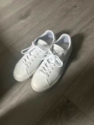 Vita Lacoste sneakers i läder - Fräscha vita sneakers från Lacoste i klassisk låg modell. Skorna är gjorda i slätt läder med vita snören och har den ikoniska gröna krokodilloggan på sidan. Perfekta för en clean och stilren look. ”LITE SMUTSIGA I BOTTEN ANNRS SOM HELT NYA”