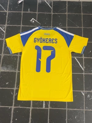 Sverige fotbollströja Gyökeres 17 - Gul och blå Sverige fotbollströja från Adidas med Gyökeres och nummer 17 på ryggen. Tröjan har korta ärmar, blå detaljer på axlar och ärmslut samt det svenska landslagets emblem på bröstet. Perfekt för match eller träning.