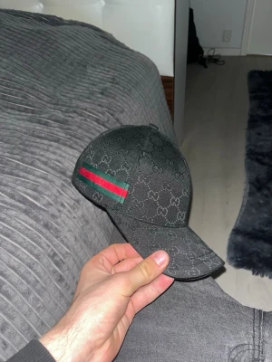 Svart Gucci keps med GG-mönster - Snygg svart keps från Gucci med klassiskt GG-monogram över hela kepsen. På sidan finns den ikoniska gröna och röda randen. Kepsen har böjd skärm och justerbar passform. Perfekt accessoar för att lyfta din streetwear-look.