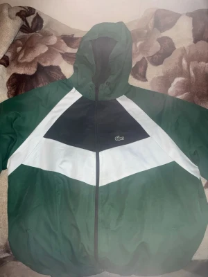 Grön och svart vindjacka från Lacoste - Snygg vindjacka från Lacoste i grönt, svart och vitt med ikoniska krokodilloggan på bröstet. Jackan har huva, dragkedja och sportig design med färgblock. Perfekt för en streetwear-look och skyddar mot vind. Materialet är lätt och känns smidigt. Passar XL jätte bra