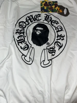 Vit Chrome Hearts x Bape t-shirt - Vit t-shirt från Bape i samarbete med Chrome Hearts. Stor svart logga framtill med klassisk Bape-apa i mitten och Chrome Hearts-typografi runtom. T-shirten har rund hals och korta ärmar. Perfekt för dig som gillar streetwear och unika samarbeten.