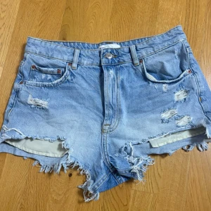 Ljusa slitna jeansshorts med fransar - Säljer ett par ljusblå jeansshorts med rejält slitna detaljer och fransig kant. Shortsen har klassisk femficksmodell, hög midja och dragkedja med knapp. Perfekta för en avslappnad och trendig sommarstil.
