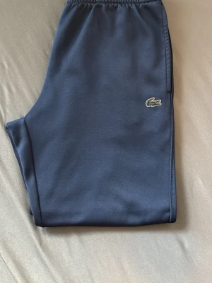 Mörkblå mjukisbyxor från Lacoste - Mörkblå mjukisbyxor från Lacoste med klassisk krokodillogga på benet. Byxorna har resår i midjan och muddar vid bensluten. Perfekta för en avslappnad och sportig stil. Materialet är mjukt och stretchigt för extra komfort.