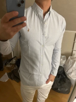 Randig skjorta från Ralph Lauren - Klassisk vit skjorta med tunna blå ränder från Ralph Lauren. Skjortan har lång ärm, knappar framtill och en broderad logga på bröstet. Snygg och tidlös passform med button-down krage.