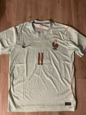 Frankrike Tredje VM-kit Fotbollströja Nike Olise  - Ljusgrön Frankrike VM fotbollströja från Nike med bronsfärgade detaljer, nummer 11 och namnet Olise på ryggen. Tröjan har korta ärmar, broderad Frankrike-logga och två stjärnor på bröstet. Tillverkad i lätt och ventilerande material med Aero-Fit-teknologi.