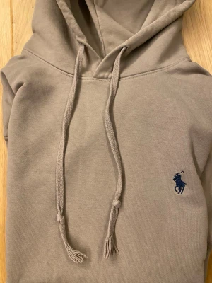 Beige hoodie från Polo Ralph Lauren - Snygg beige hoodie från Polo Ralph Lauren med klassisk huva och dragsnören. Broderad marinblå logga på bröstet och stor magficka framtill. Perfekt för en chill och stilren look. Mjuk bomullskvalitet som känns skön mot huden.