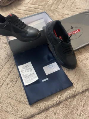 Svarta Prada sneakers i läder och mesh - Snygga svarta sneakers från Prada med ovandel i en mix av läder och mesh. Klassisk låg modell med svarta skosnören och diskret Prada-logga på plösen. Skorna har en chunky sula och stilren design som passar till många outfits.
