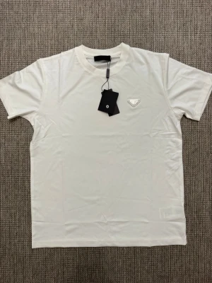 Vit t-shirt från Prada - Snygg vit t-shirt från Prada med klassisk passform och rund hals. T-shirten har en diskret logga på bröstet och är tillverkad i mjuk bomull. Perfekt för en stilren och enkel look.