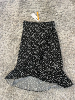 Svart blommig volangkjol Gina Tricot - Supersöt svart kjol från Gina Tricot med små vita blommor och volangkant nertill. Kjolen har omlott-look och är gjord i ett mjukt, lätt material som passar perfekt till sommaren. Den här aldrig använd och prislappen sitter kvar.